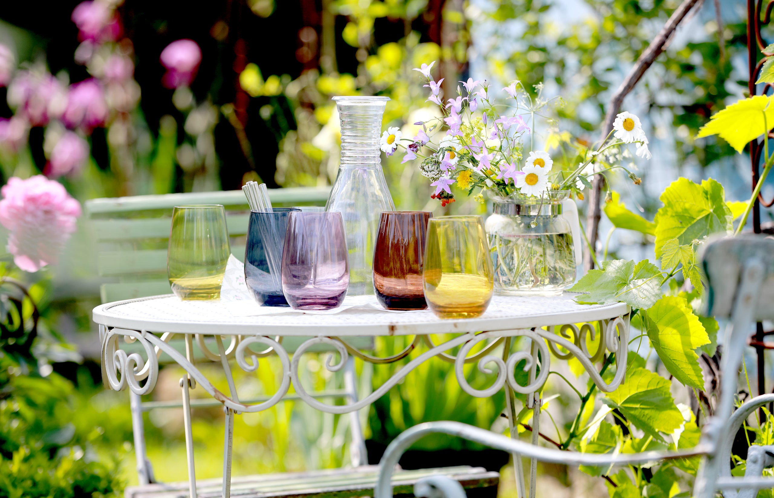 Bunte Gläser Set in verschiedenen Farben auf einem Gartentisch mit einer Blumenvase und dekorativem Hintergrund
