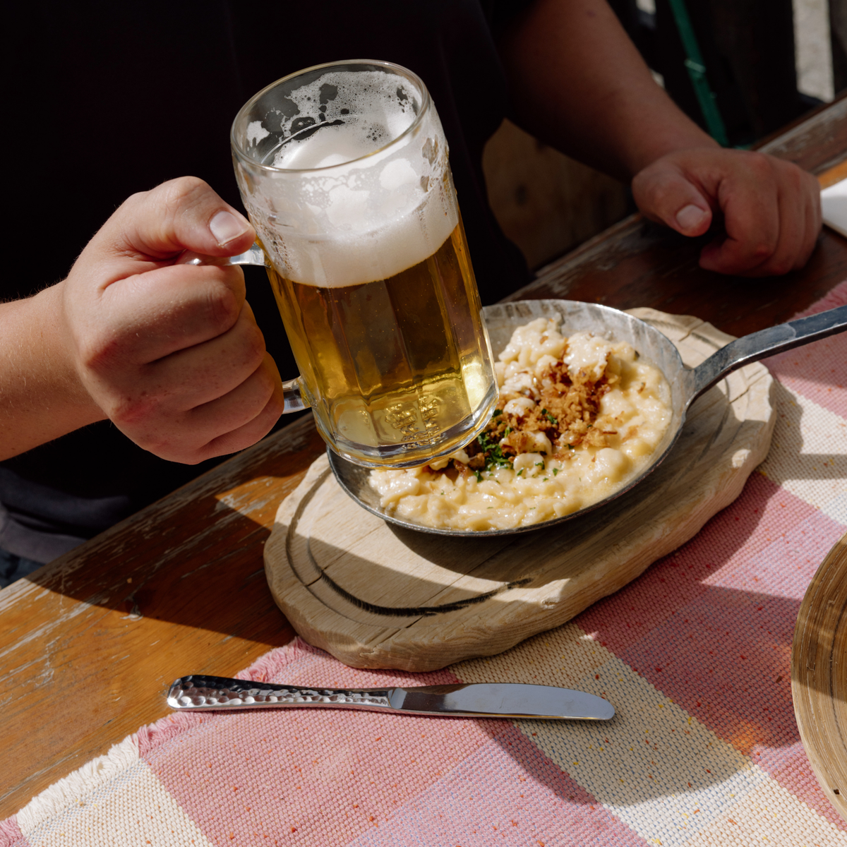 Oberglas Ecken Bierkrug, 500ml, gefüllt mit Bier in einer männlichen Hand in hebender Bewegung am Tisch über einem Teller Käsenudeln